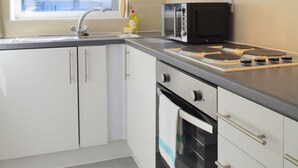 A compact kitchen with white cabinets, a black microwave, a stainless steel sink, and a window with a black blind. - The London Agent Fitzrovia Studio 3 (London)