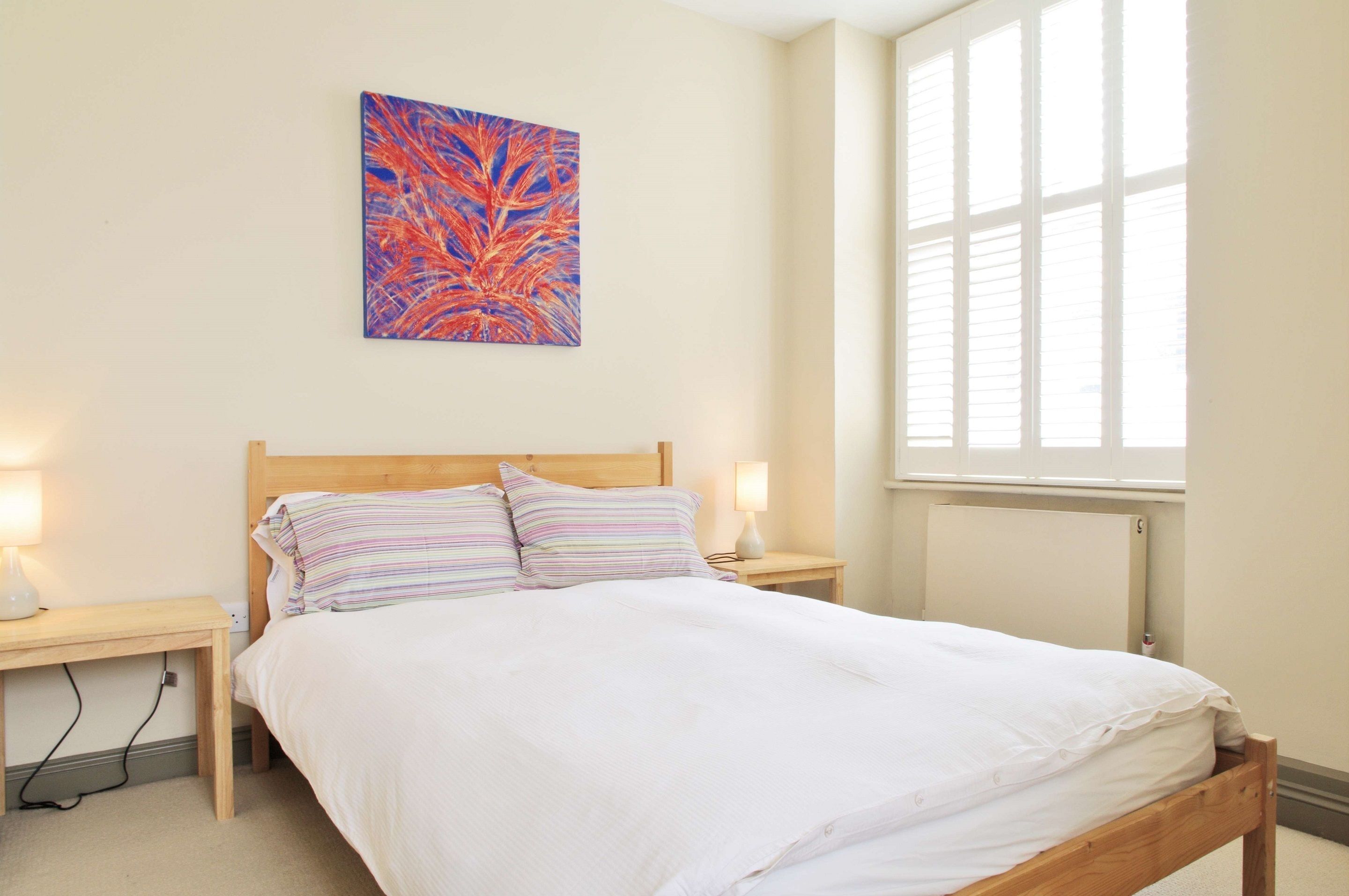 Une chambre à coucher avec un lit en bois, deux lampes de chevet, une table de nuit, une fenêtre avec des stores et un tableau abstrait aux couleurs vives accroché au mur.