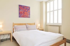 Ein Schlafzimmer mit einem hölzernen Bett, zwei Nachttischlampen, einem Nachttisch, einem Fenster mit Jalousien und einem farbenfrohen abstrakten Gemälde an der Wand.