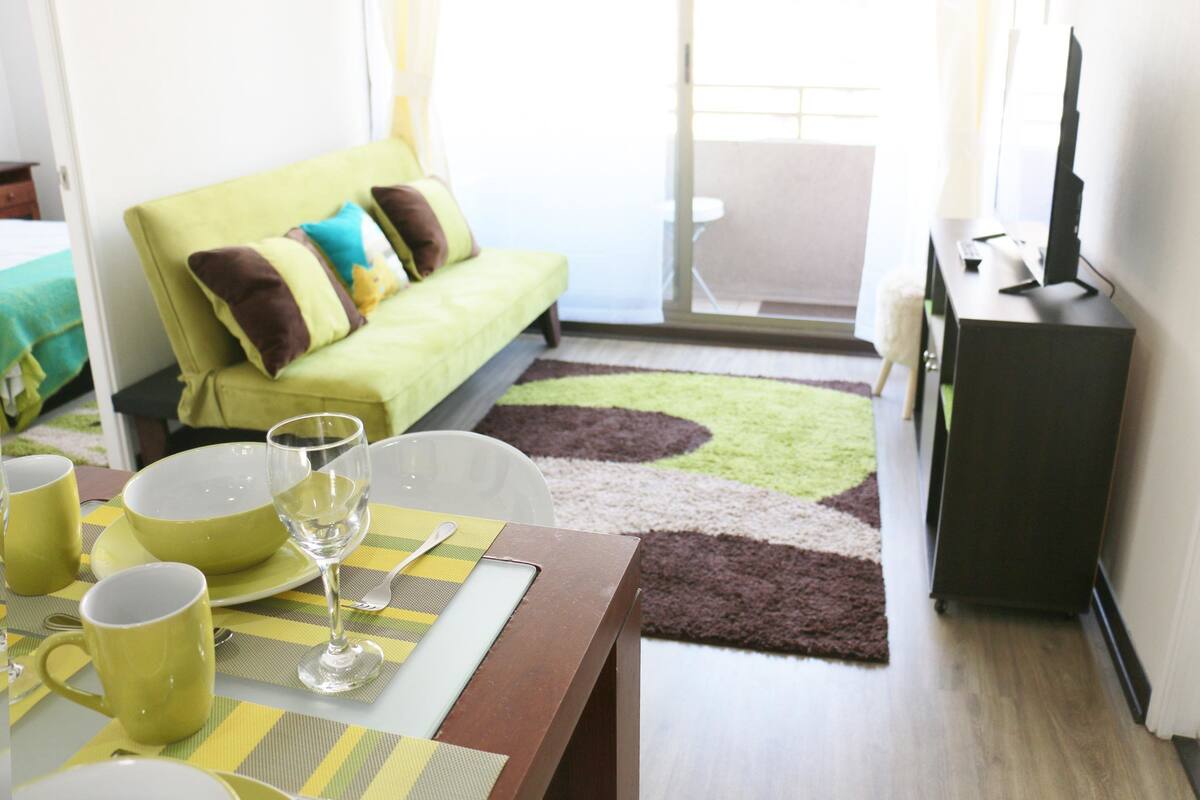 A modern living room with a green sofa, a dining table with yellow placemats, and a flat-screen TV on a stand.