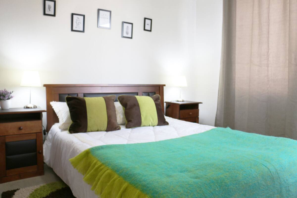A neatly made bed with a white duvet and a teal blanket, flanked by two bedside tables with lamps, a wooden nightstand, and a small potted plant.