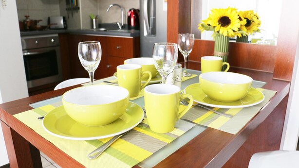 A dining table set with yellow bowls and cups, a vase with sunflowers, and a kitchen in the background. - Chile4Rent Eleuterio Ramirez (Santiago)