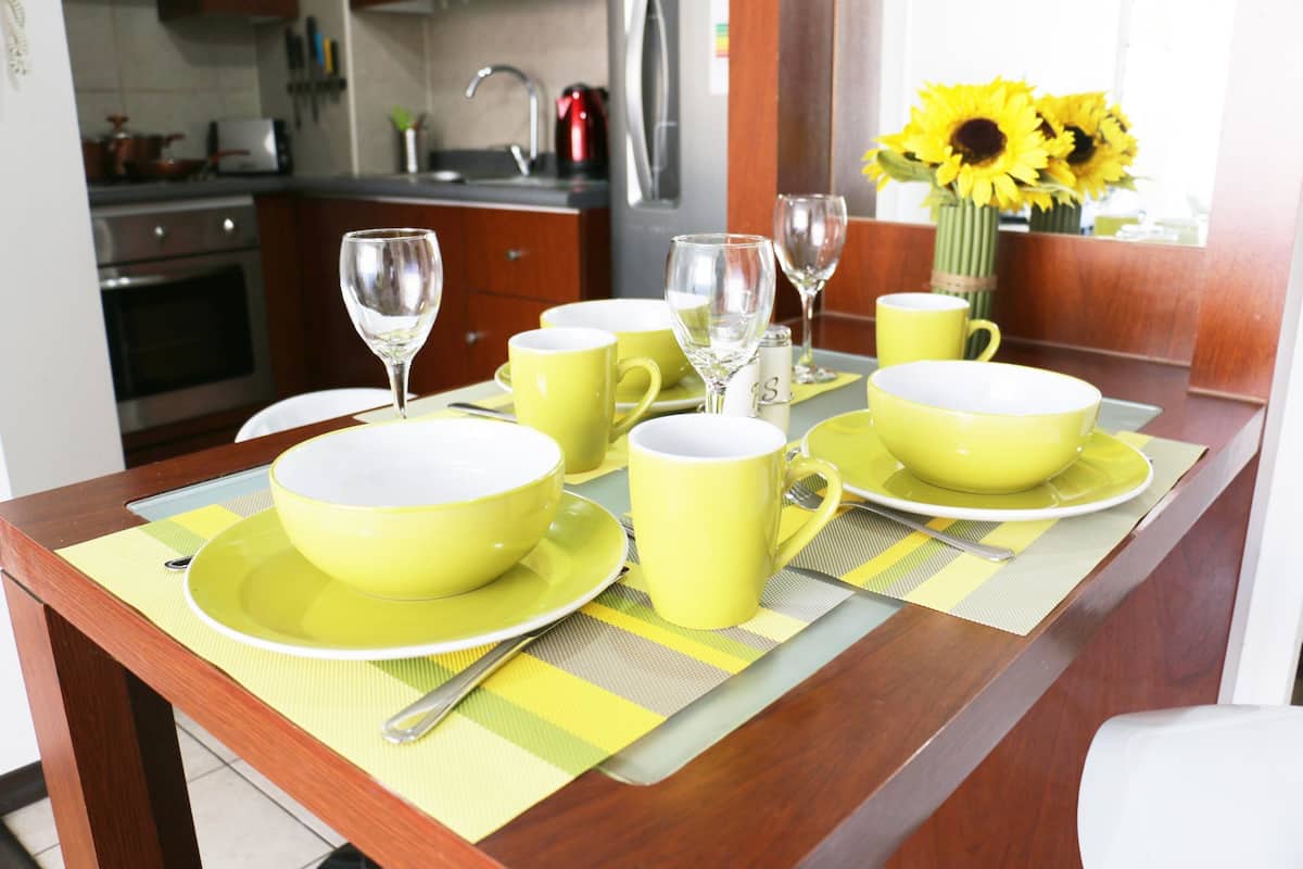 A dining table set with yellow bowls and cups, a vase with sunflowers, and a kitchen in the background.