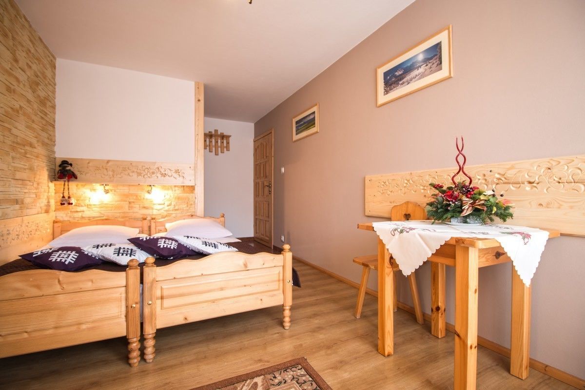 Ein hölzernes Bett mit Kopfteil, ein kleiner Holztisch mit weißer Tischdecke und ein an der Wand befestigtes Regal mit einem dekorativen Gegenstand.