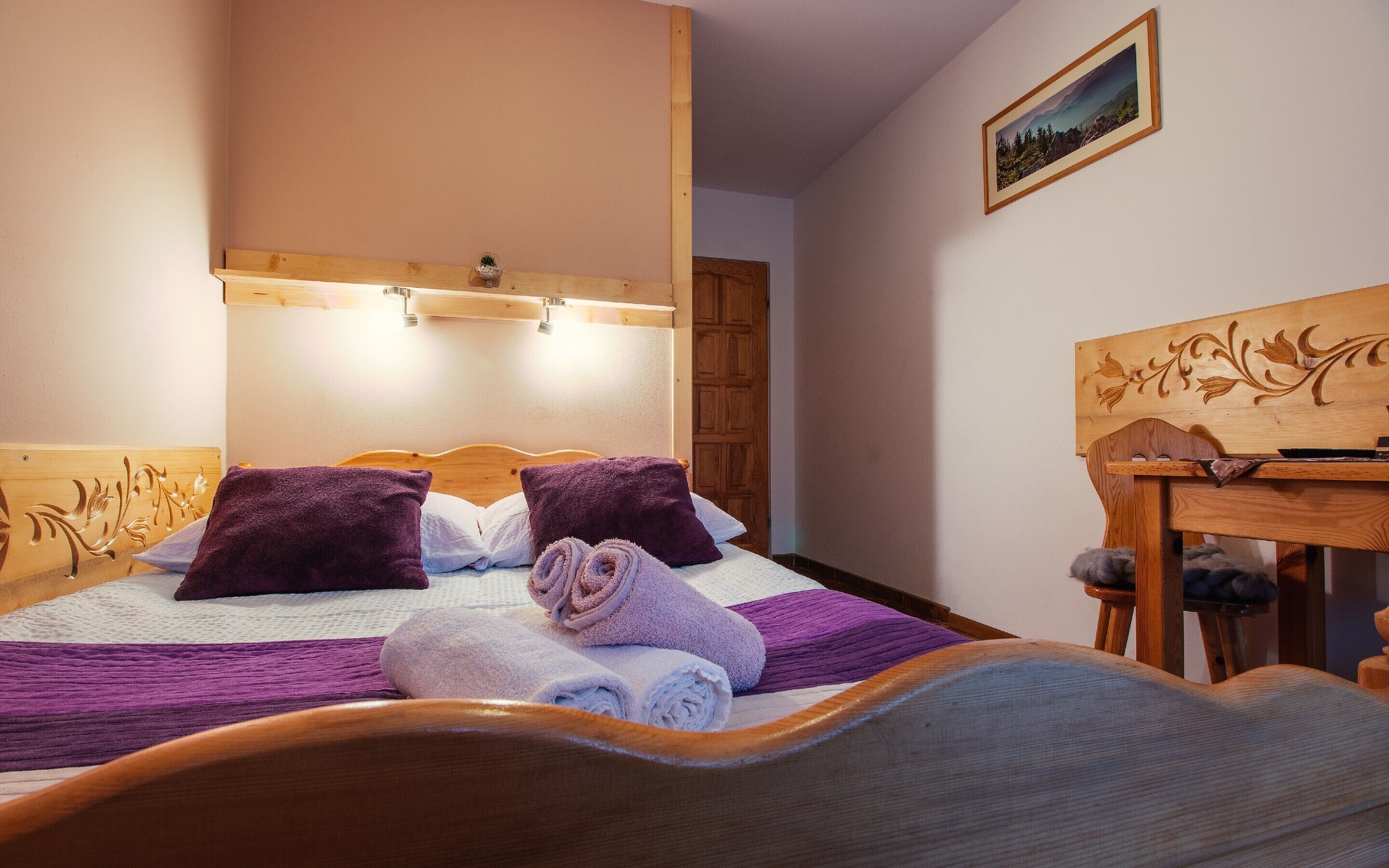 A neatly made bed with purple and white towels, a wooden headboard with carved designs, a wooden table with a tray, and a framed picture on the wall.