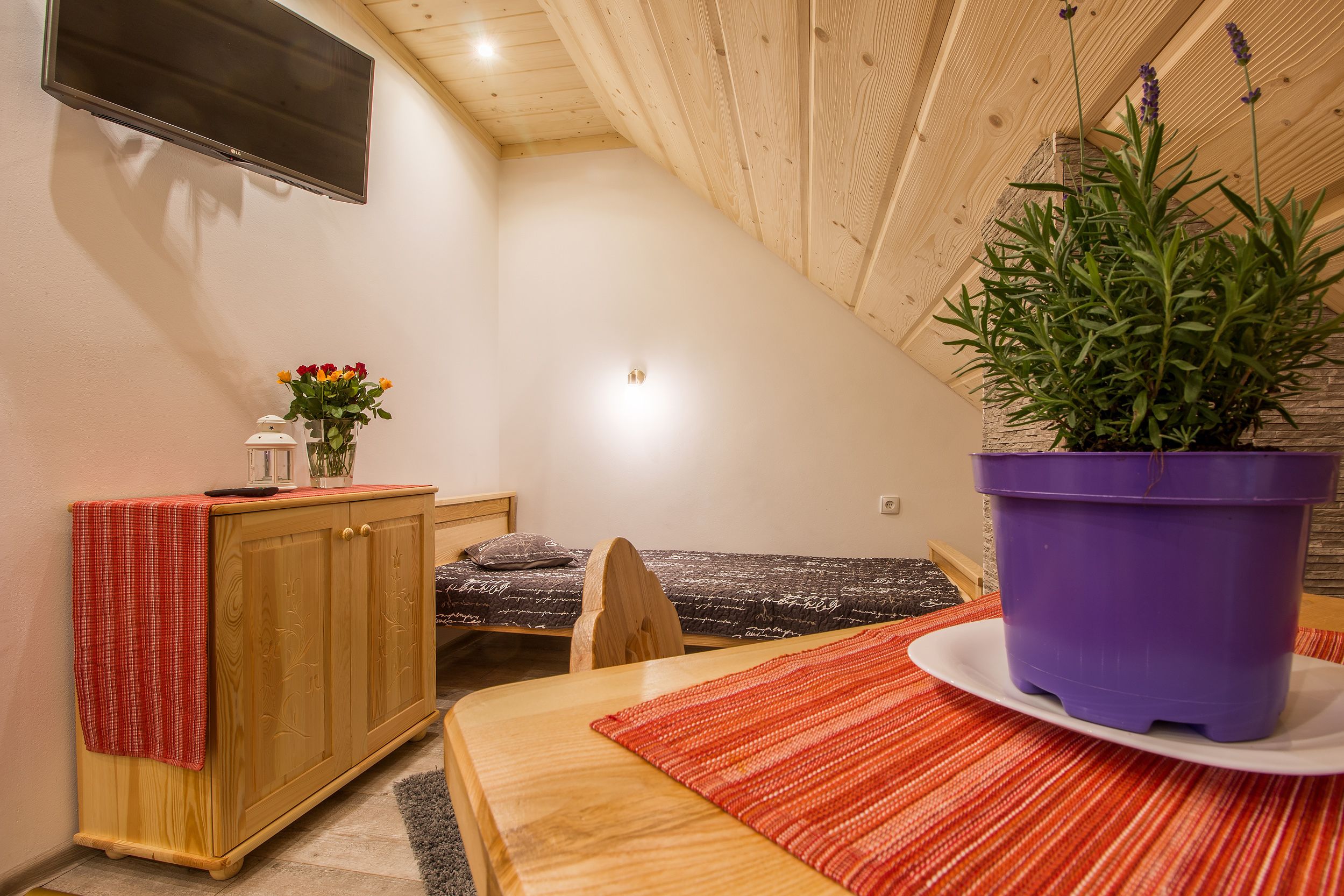 A small, cozy room with a wooden ceiling, a bed, a desk with a plant, and a TV mounted on the wall.
