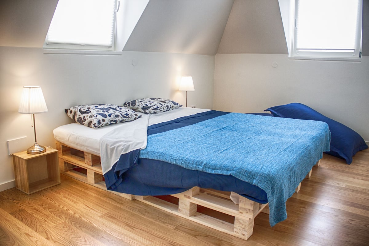 A loft bedroom with a sloped ceiling, wooden flooring, a bed with blue bedding, two bedside lamps, and a skylight.