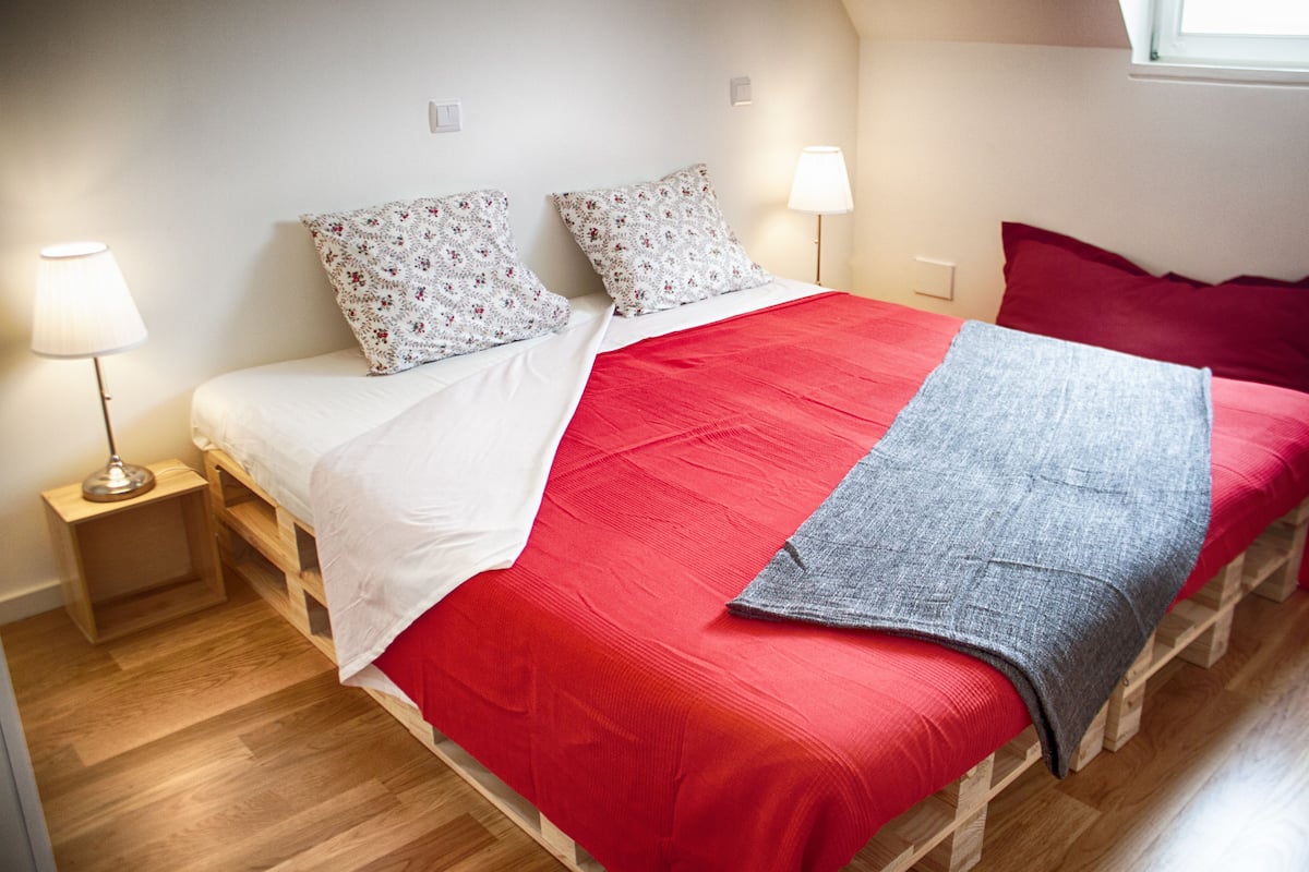 A bed with a red and white bedspread, two pillows, a grey throw, and a bedside lamp.
