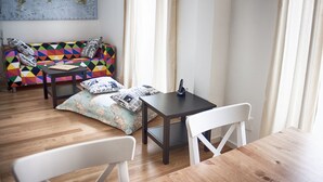 A room with a wooden floor, a white chair, a small table, a colorful sofa with patterned cushions, and a world map on the wall.