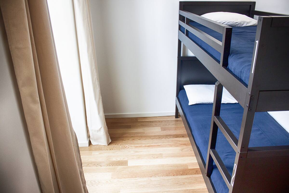 A bunk bed with a blue mattress and white pillows, wooden flooring, and a curtain partially drawn.