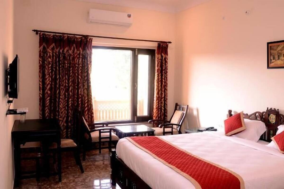 Quarto de hotel com cama, escrivaninha, cadeira, televisão e janela com cortinas.