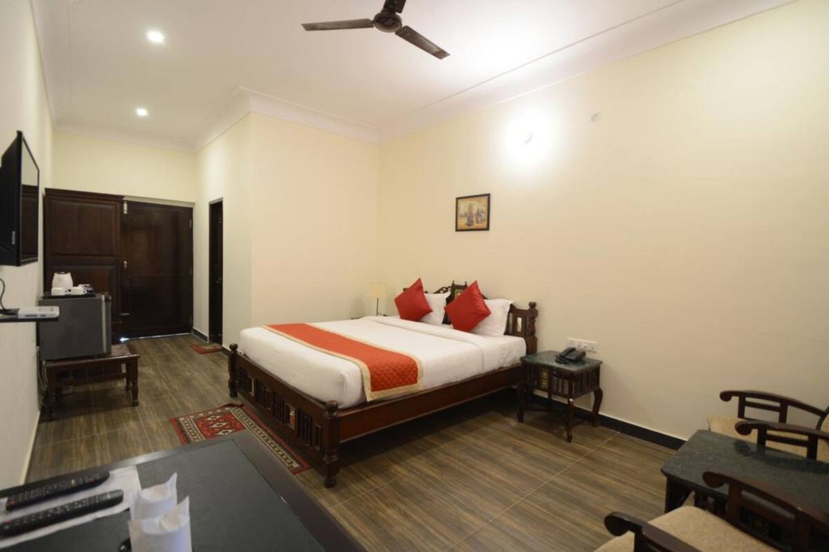 Quarto de hotel com cama, escrivaninha, cadeira, luminária de mesa, telefone e ventoinha no teto.