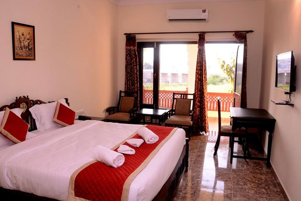 Quarto de hotel com cama, escrivaninha, cadeira, TV e varanda com cortinas.