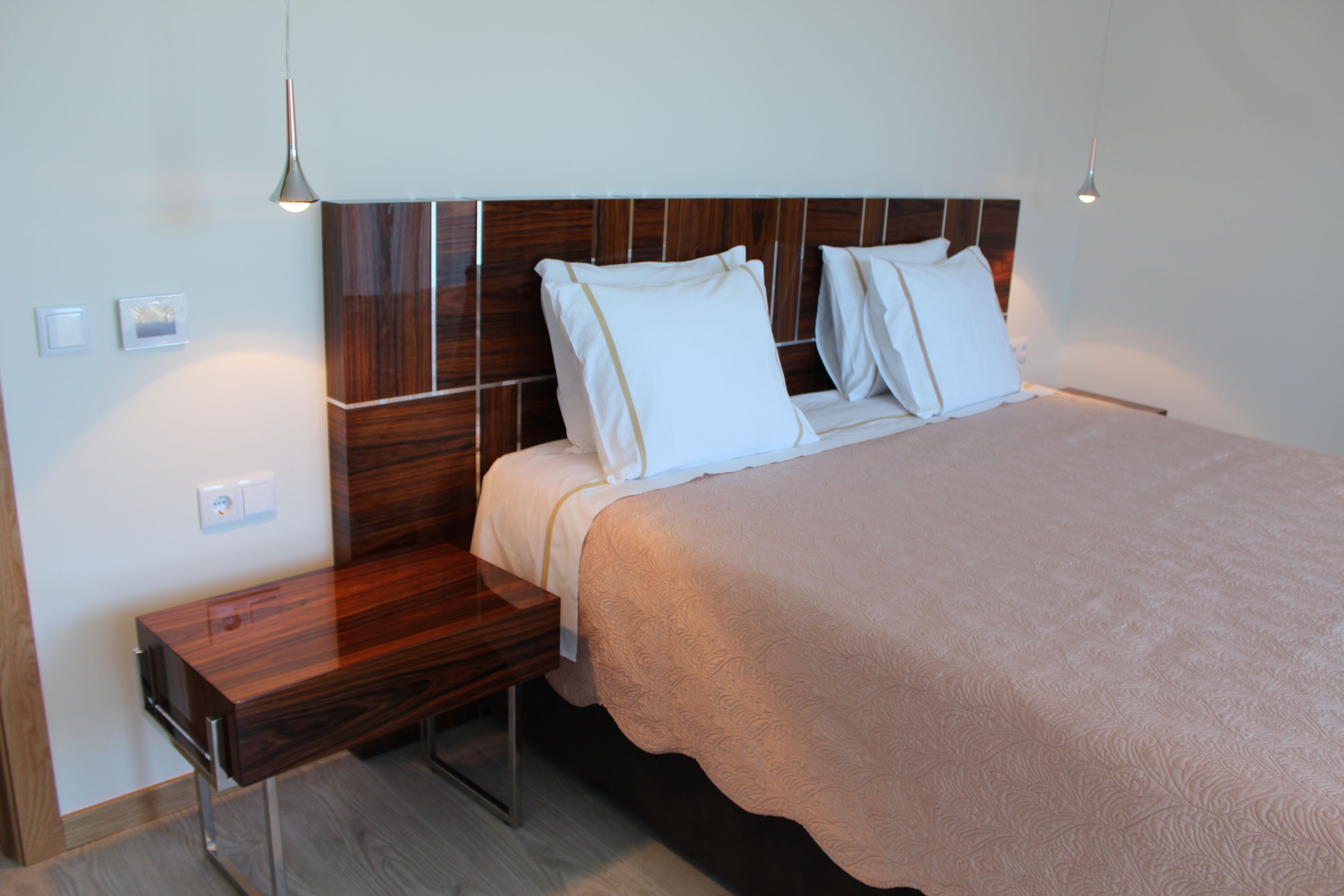 Ett modernt hotellrum med en sänggavel i trä, ett nattduksbord i trä, en säng med ett beige sängöverkast och två hänglampor.
