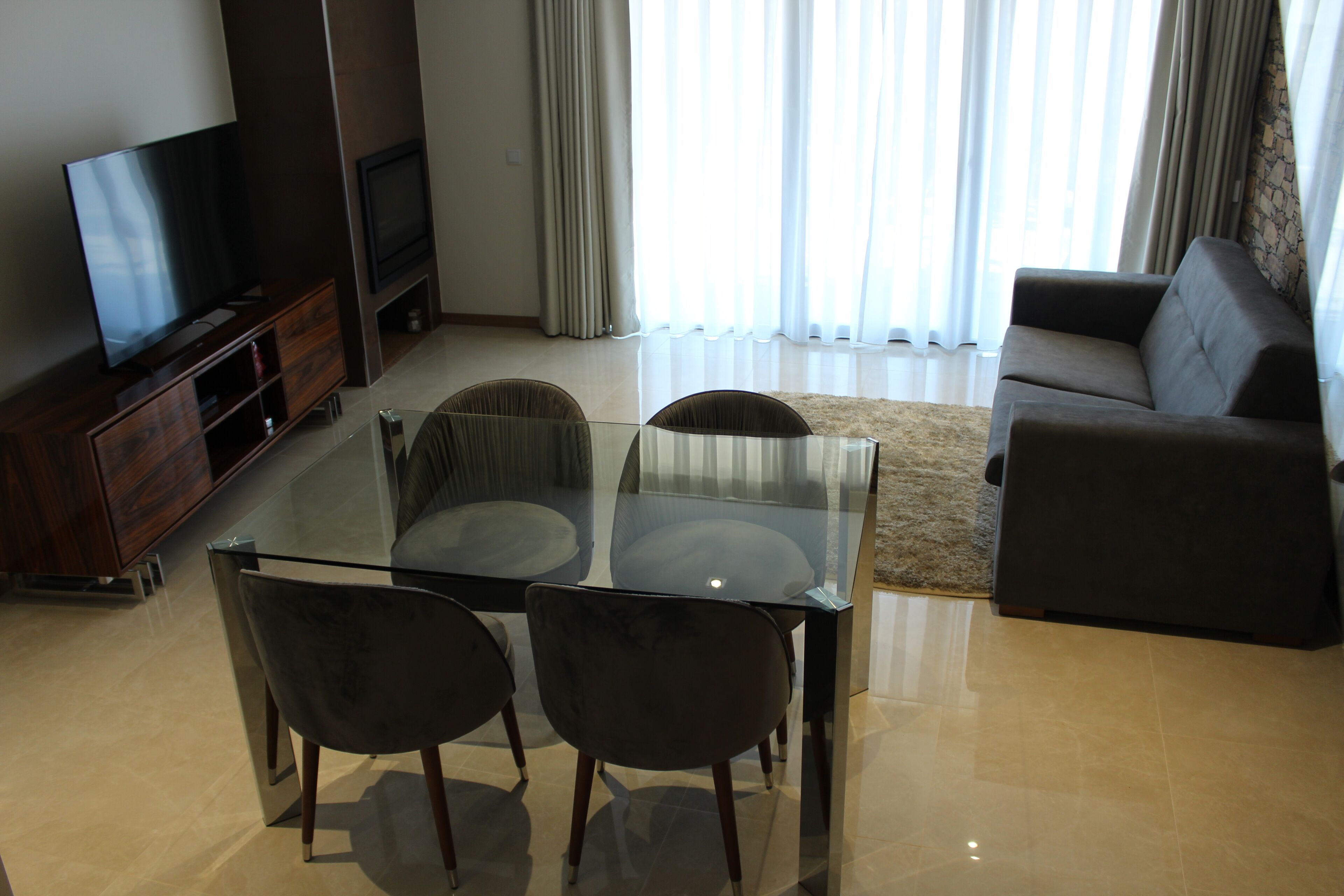 Una sala moderna con una mesa de vidrio para comer, dos sillas, un sofá, un televisor sobre un mueble de madera y un amplio ventanal con cortinas.