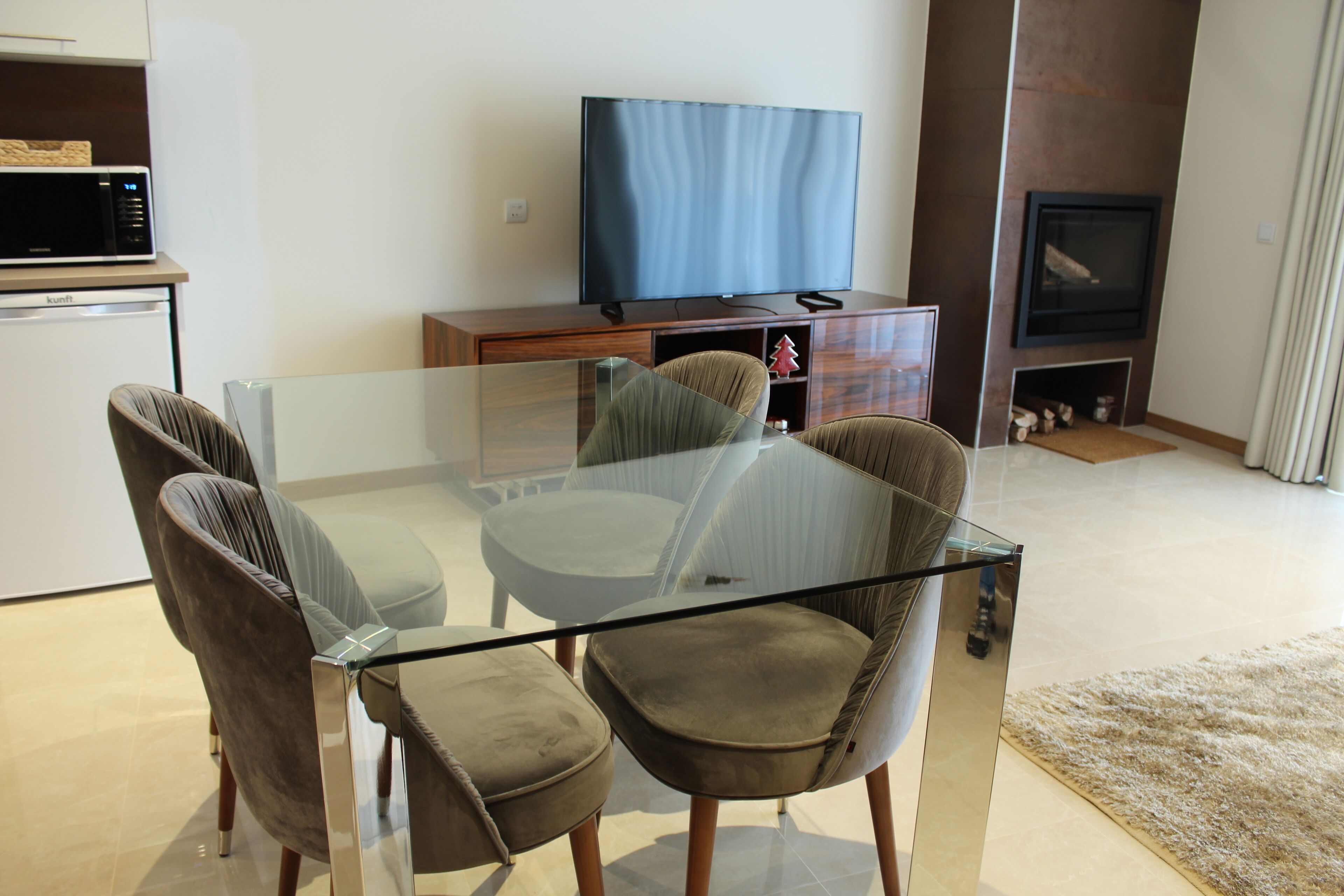 A modern living room with a glass-top dining table, cushioned chairs, a flat-screen TV, a wooden cabinet, and a fireplace.
