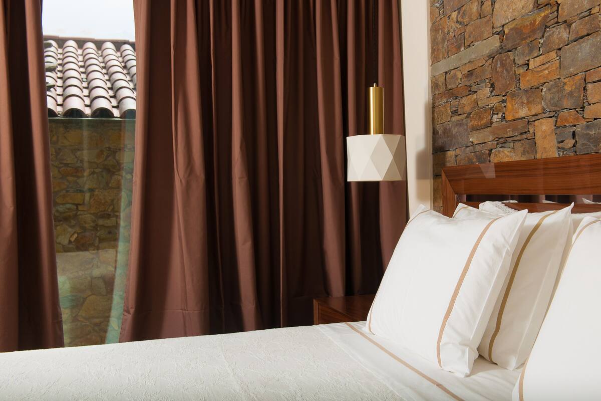 A hotel room with a bed, curtains, a stone wall, and a view of a tiled roof.