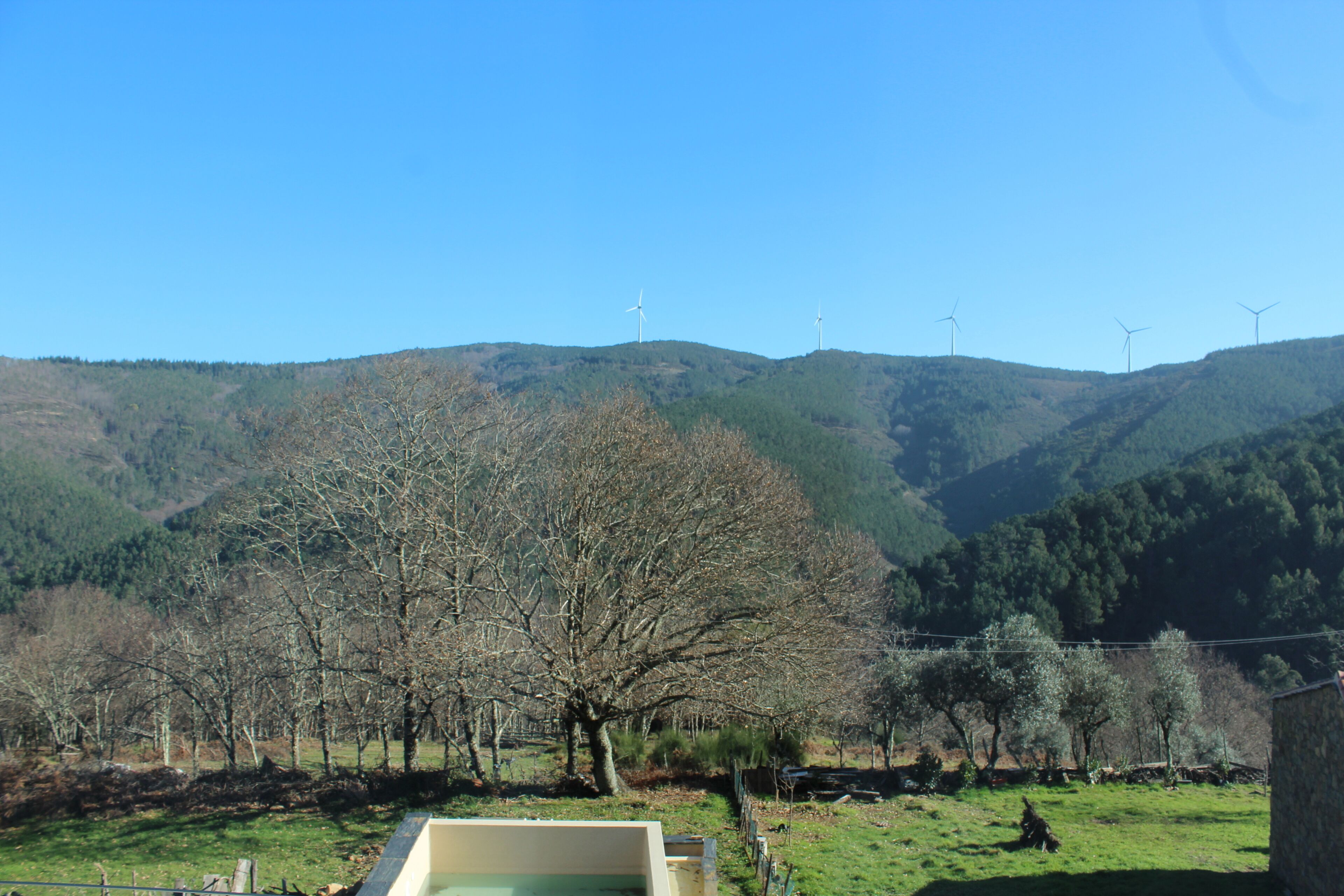 Paisaje rural con aerogeneradores en las colinas, una zona vallada con árboles y una construcción de piedra.