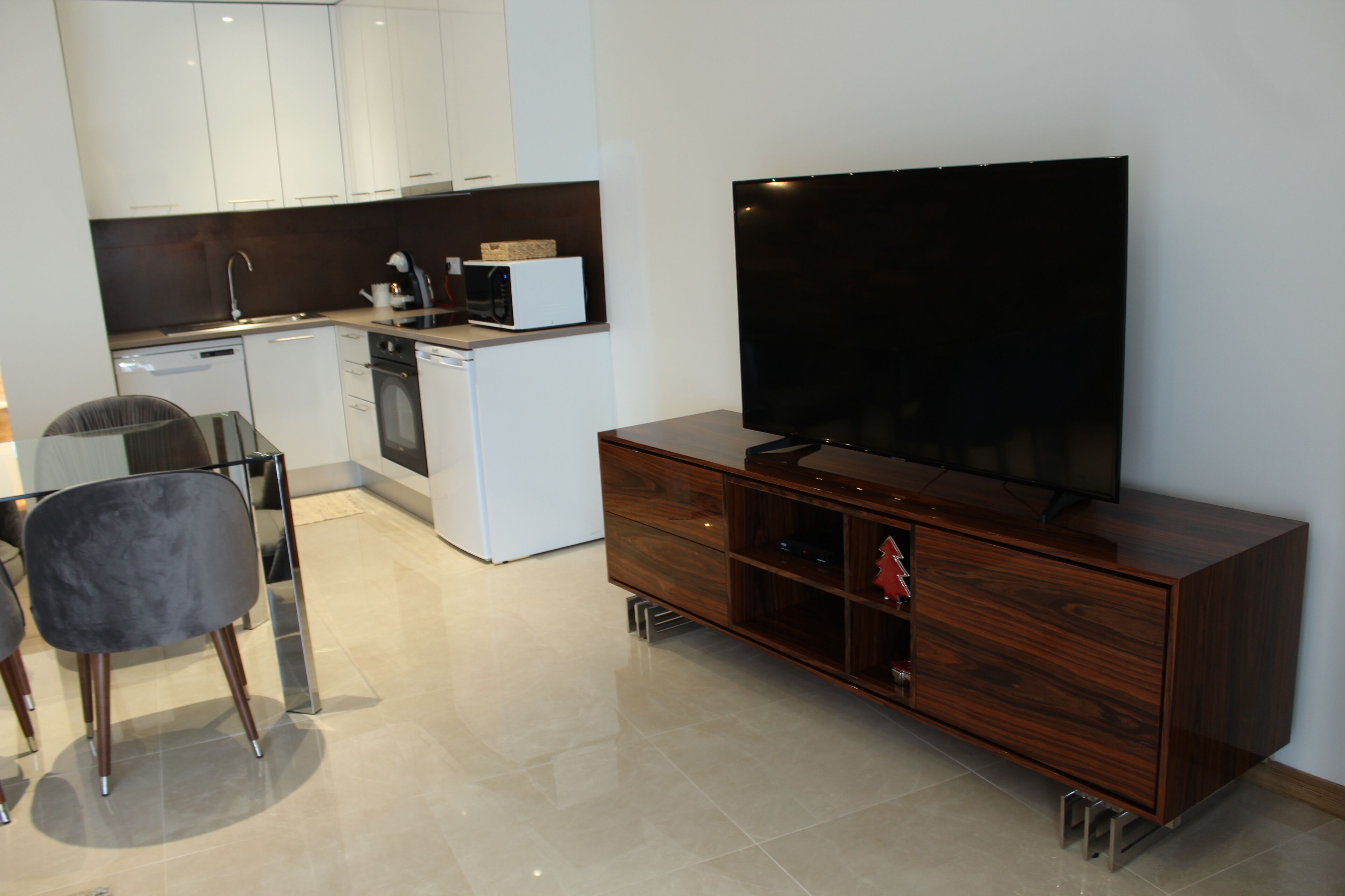 A modern kitchen with white cabinets, a microwave, and a dishwasher. There is a large flat-screen TV on a wooden stand, a dining table with chairs, and a small kitchen island with a sink.