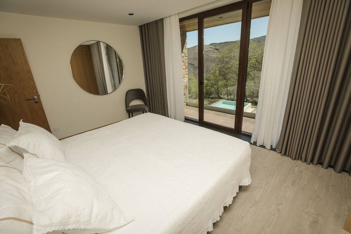 A hotel room with a large bed, a round mirror, a chair, and a view of a mountainous landscape through a sliding glass door.