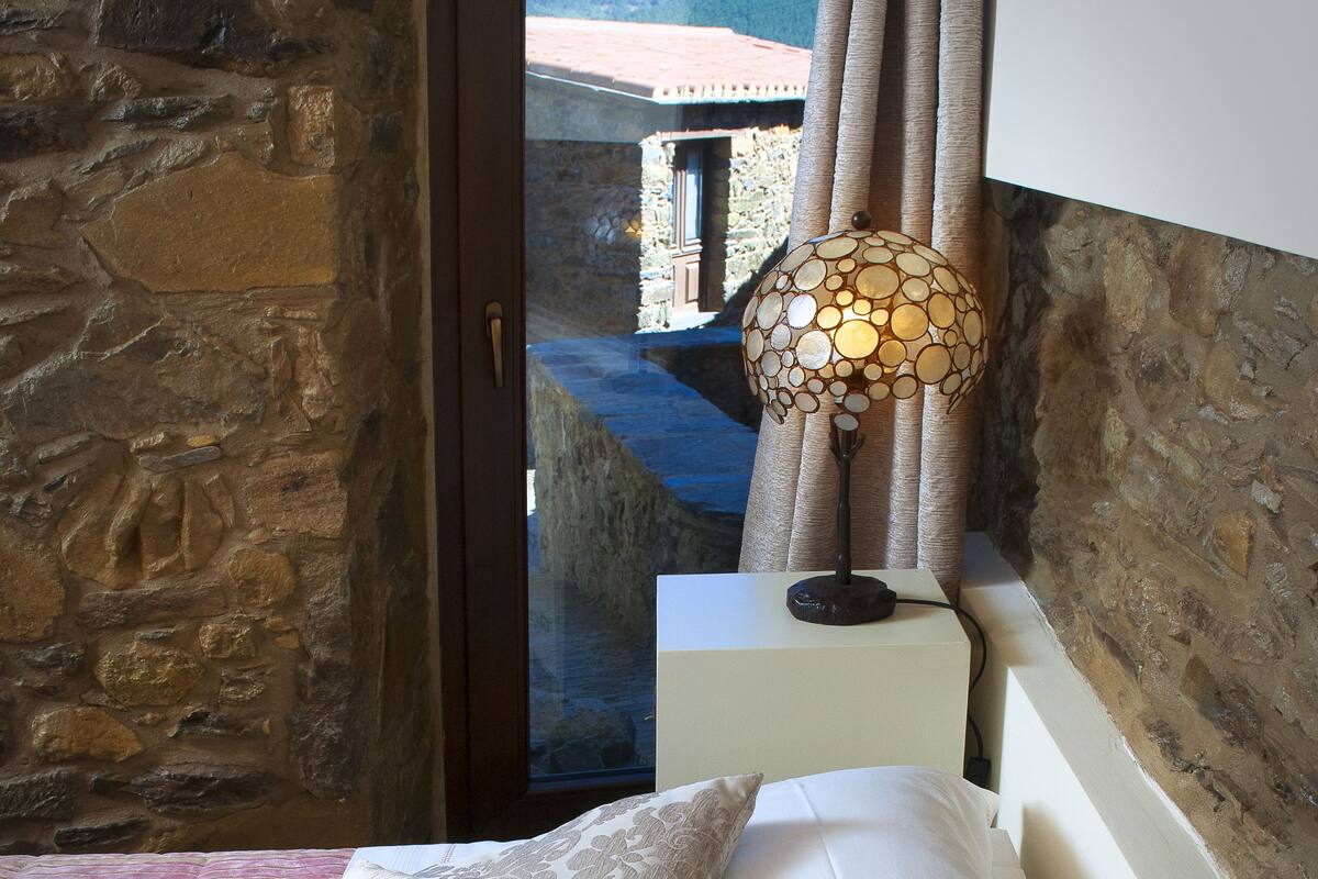 A bedroom with a stone wall, a bed with white linens, a lamp, and a view of a building through a glass door.