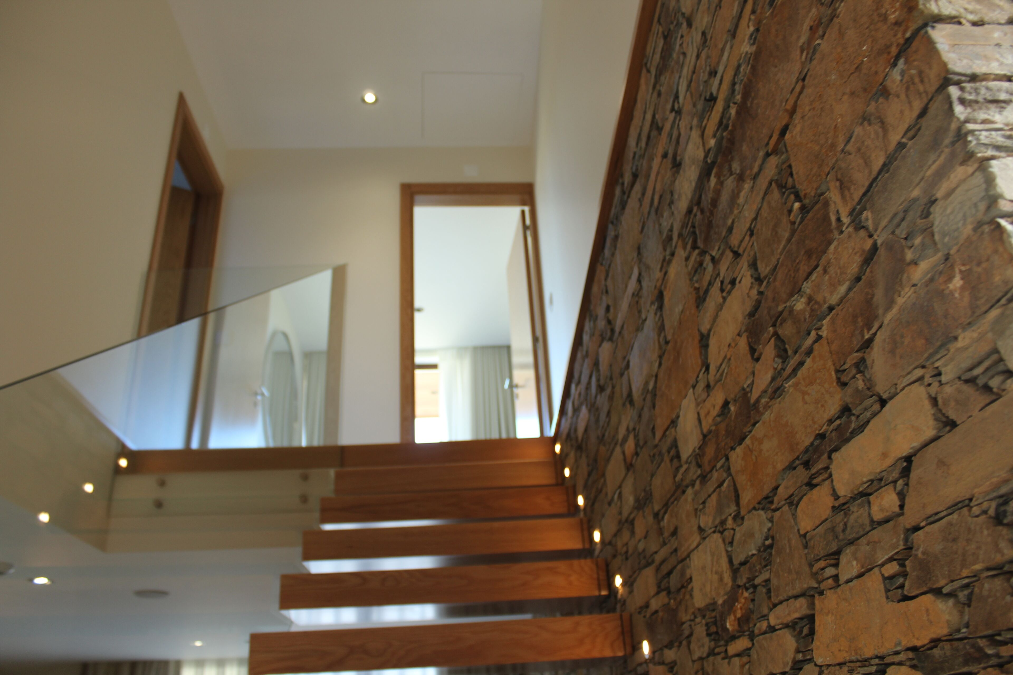 A modern staircase with wooden steps and glass railings, leading to a room with a stone wall and a door in the background.