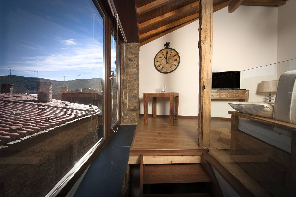 A room with a large window, wooden flooring, a stone wall, a wooden table, a clock, and a television.