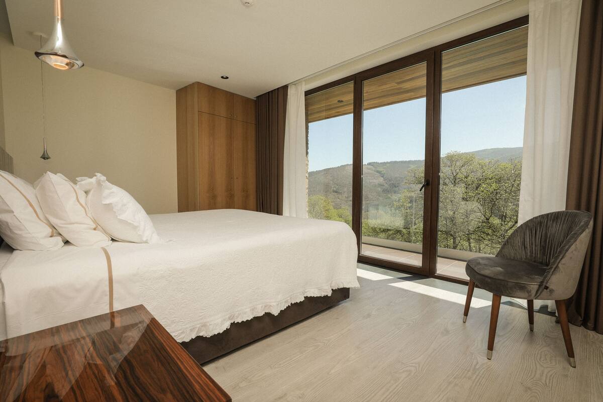 A bedroom with a large bed, a wooden wardrobe, a chair, and a view of the outdoors through a large window.