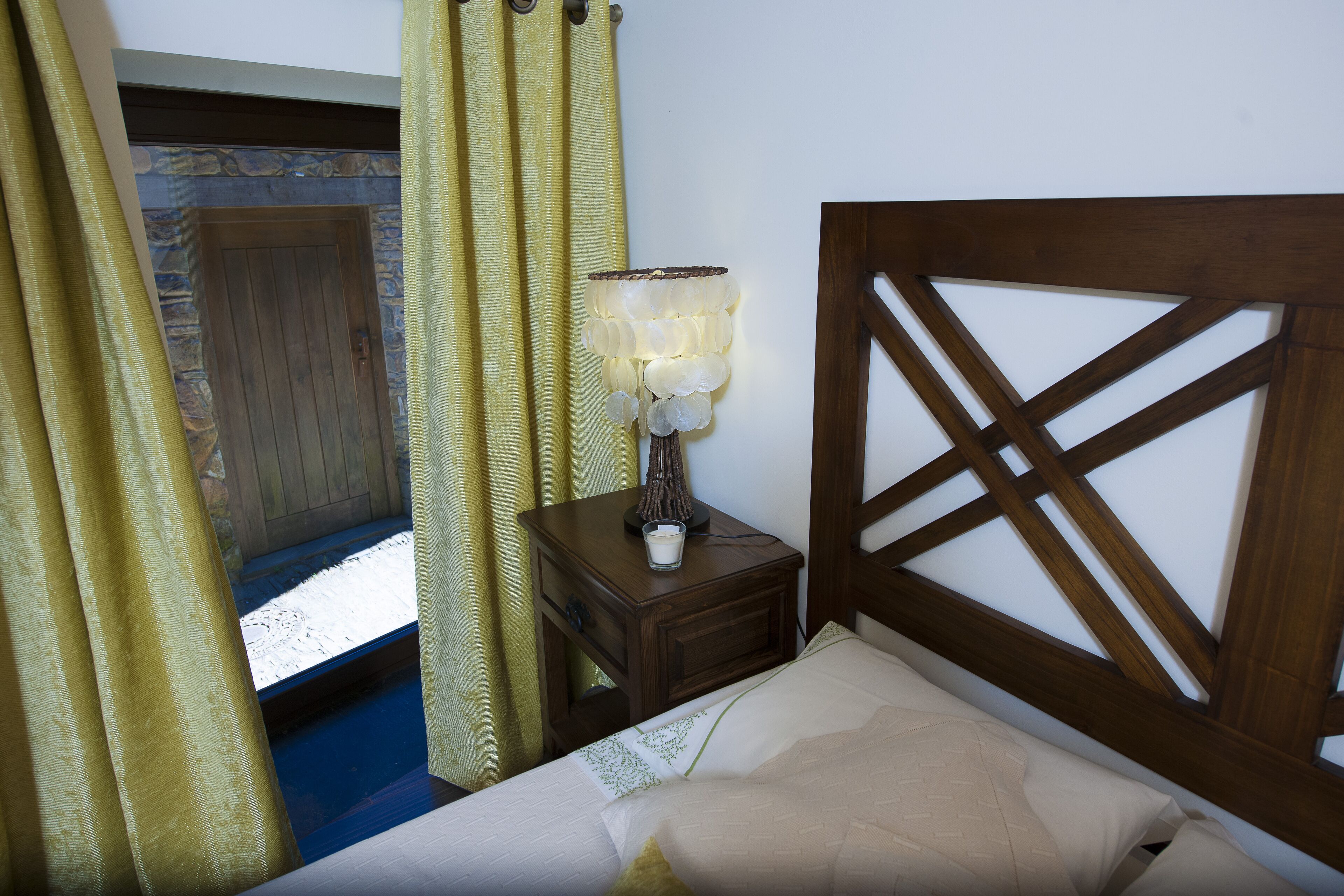 A bedroom with a wooden headboard, a bedside table with a lamp, a window with curtains, and a stone wall.