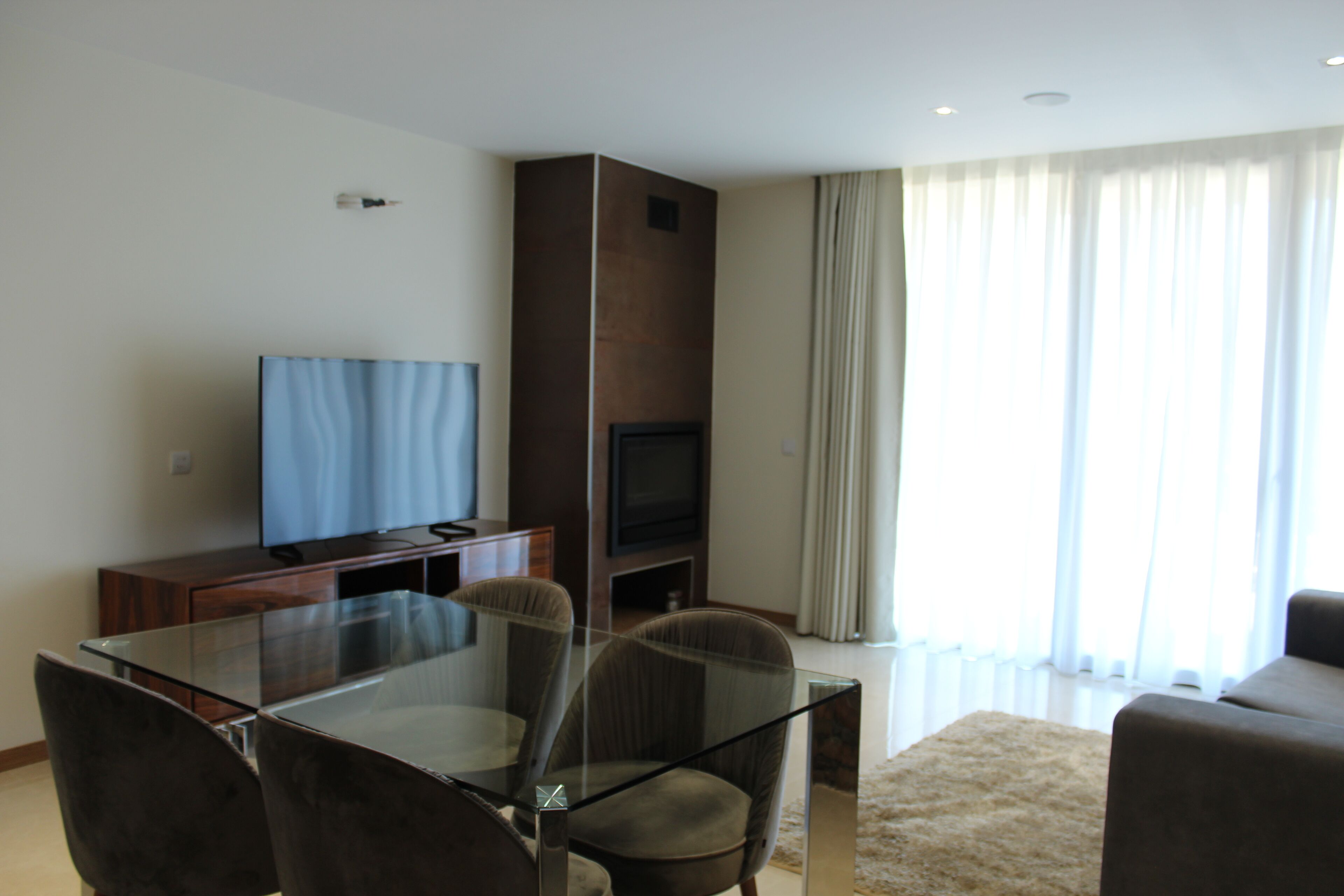 A modern living room with a glass dining table, comfortable seating, a flat-screen TV, and a large window with curtains.