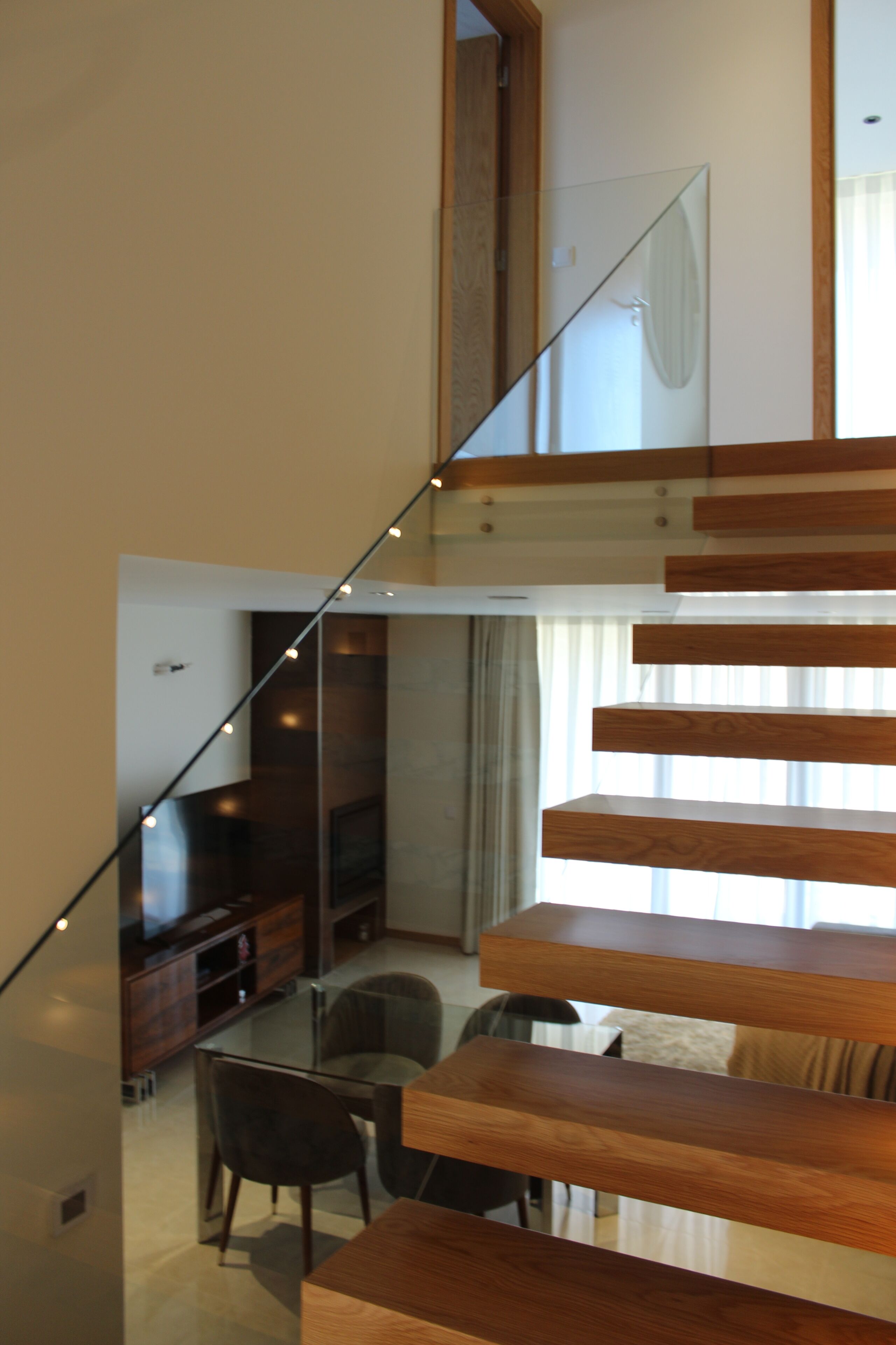 A modern staircase with wooden steps and glass railings, leading to a living area with a flat-screen TV, a sofa, and a dining table with chairs.