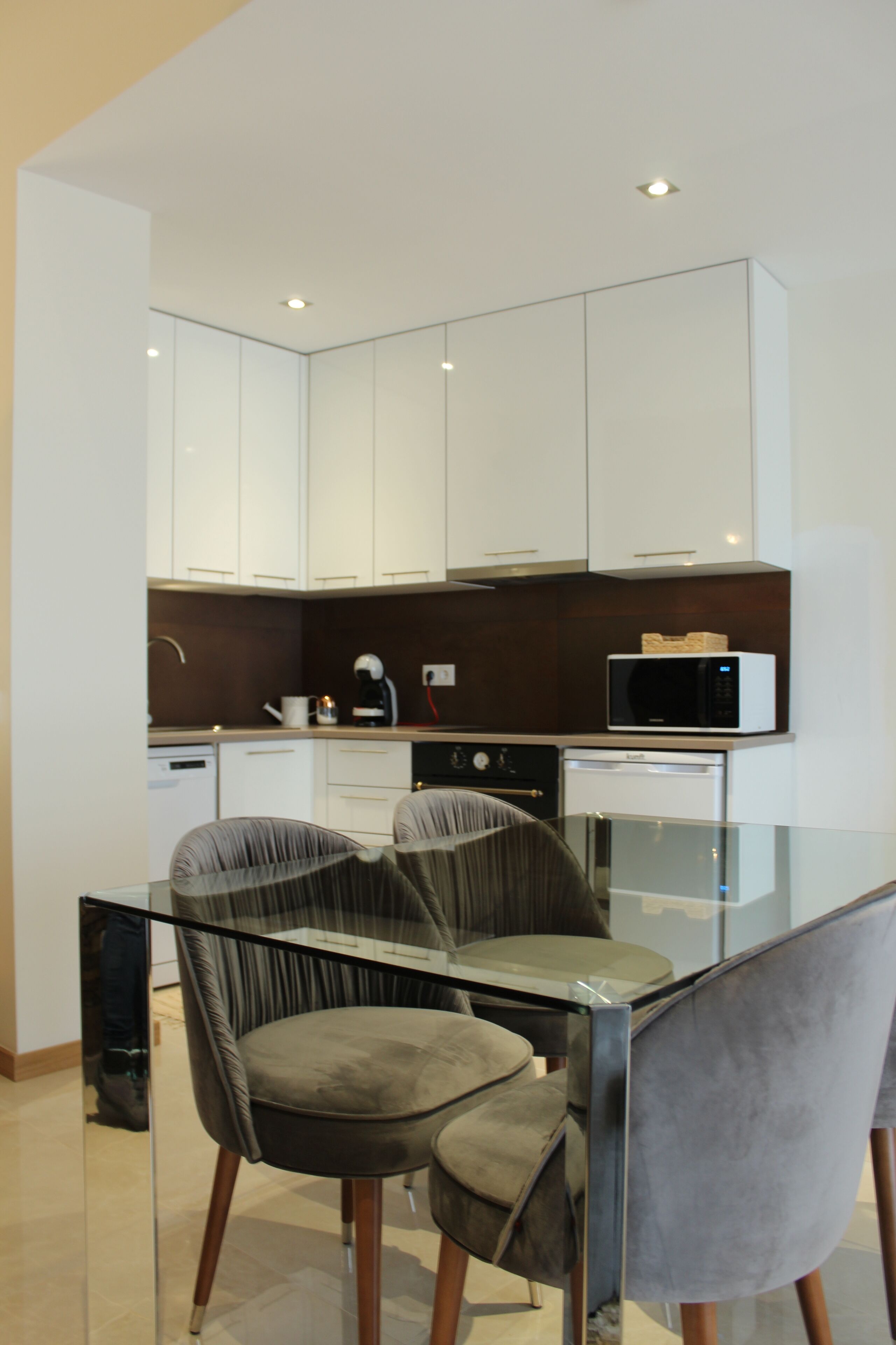 Una cocina moderna con armarios blancos, microondas y cafetera. Hay una mesa de vidrio con dos sillas.
