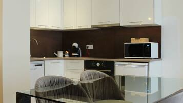 Una cocina moderna con armarios blancos, microondas y cafetera. Hay una mesa de vidrio con dos sillas.