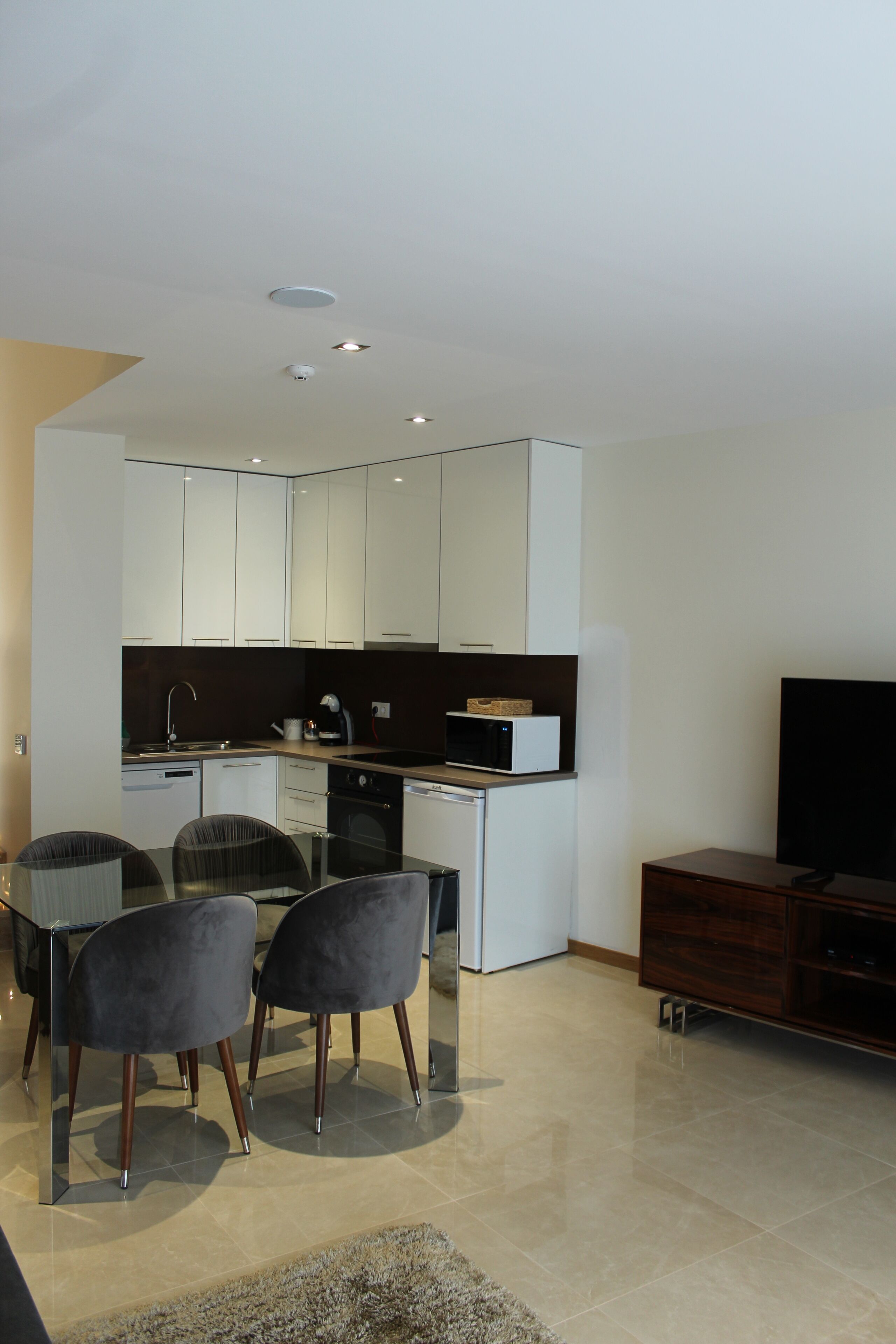 A modern kitchen with white cabinets, a microwave, and a sink. There is a dining area with a table and chairs, and a flat-screen TV on a wooden stand.