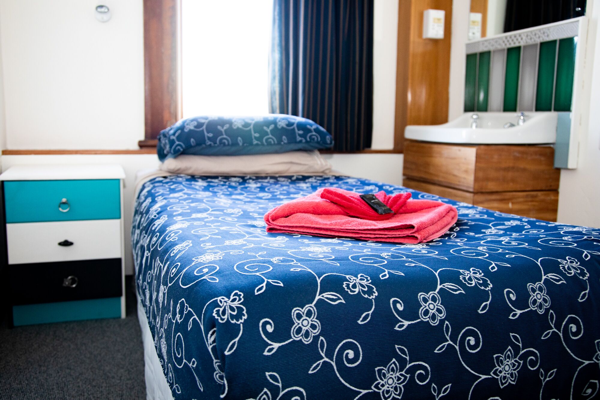 Una cama bien hecha con una colcha estampada con motivos florales, una almohada azul y una toalla roja doblada.