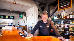 A bar area with a polished wooden counter, a bartender, and a variety of bottles and glasses. - The Brown Pub (Methven)