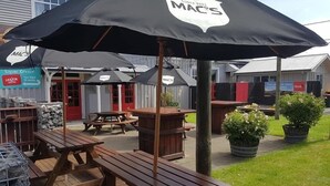 An outdoor seating area with wooden tables and picnic-style benches, covered by black umbrellas with the logo "MACS". - The Brown Pub (Methven)