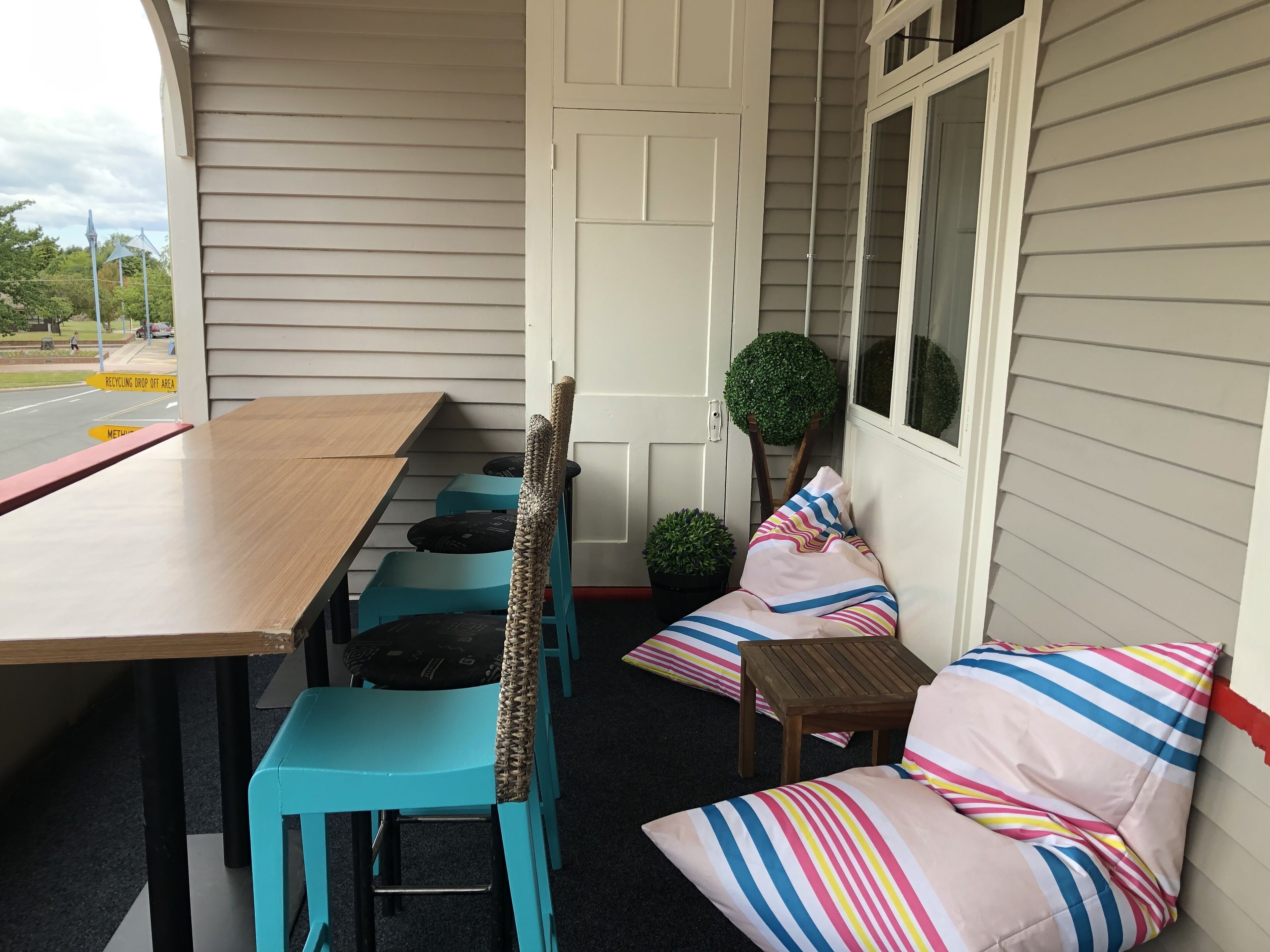 A covered patio with a wooden table and turquoise chairs, a small wooden table, and a potted plant.