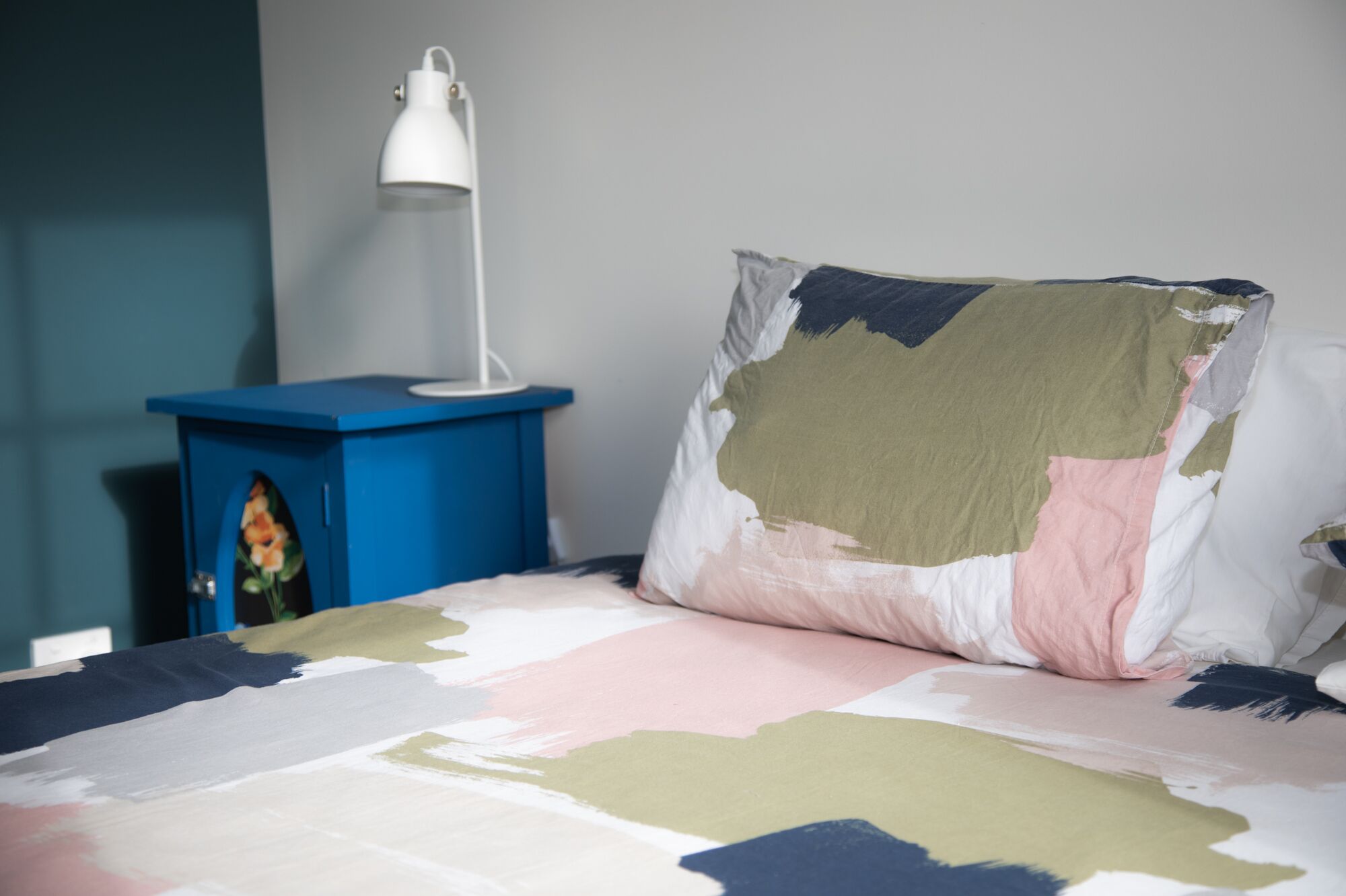 Una cama con un edredón colorido, una mesita de noche azul con una lámpara blanca y un objeto decorativo sobre la mesita.