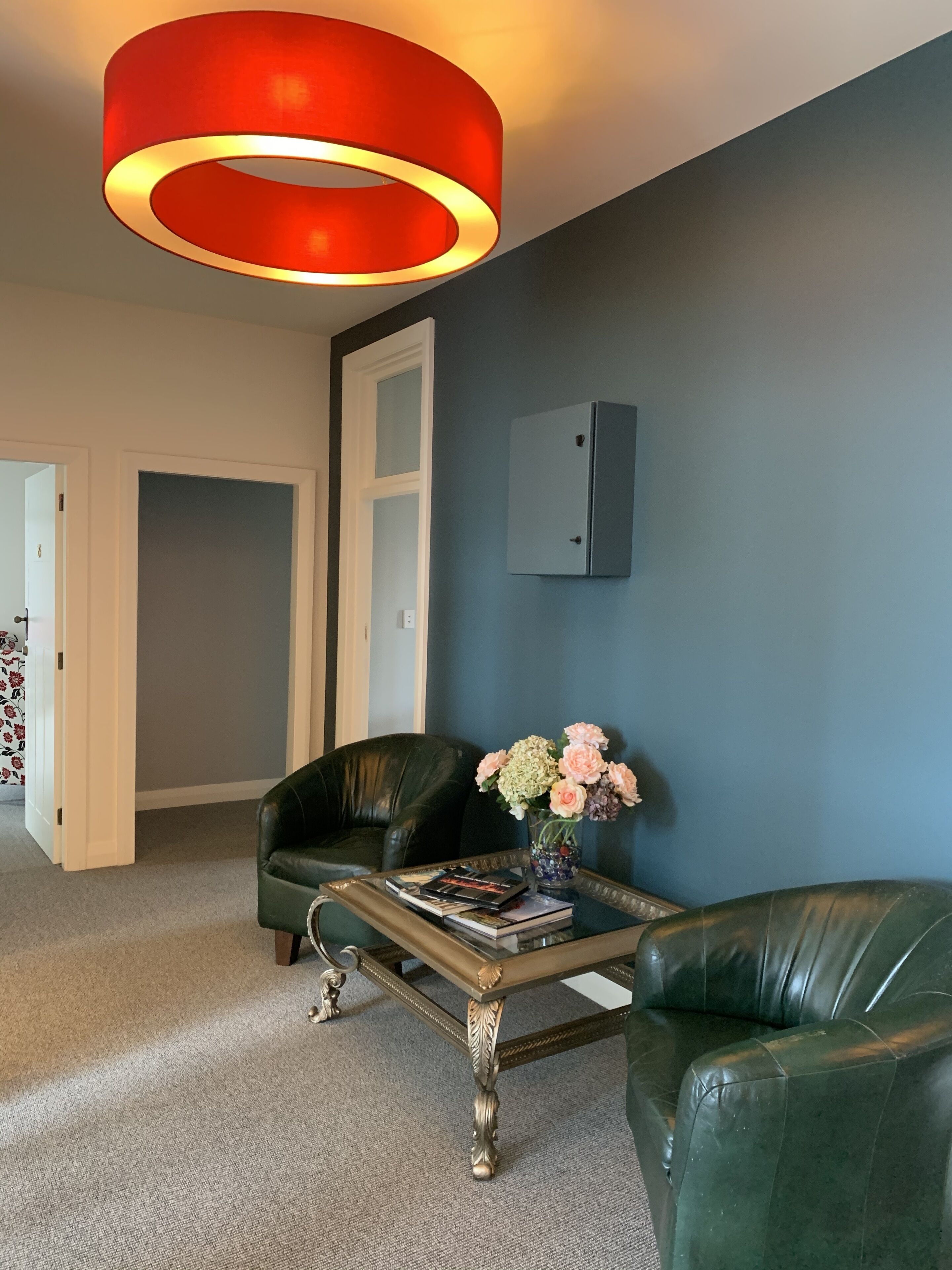 A modern living room with a dark blue wall, a red circular light fixture, a black leather sofa, a glass coffee table with books, and a vase with flowers.