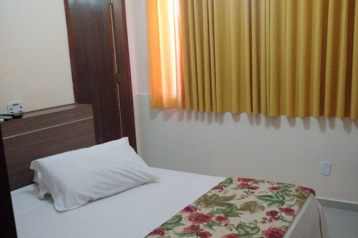 Una habitación de hotel con una cama, cabecera de madera, mesita de noche, teléfono y ventana con cortinas amarillas.