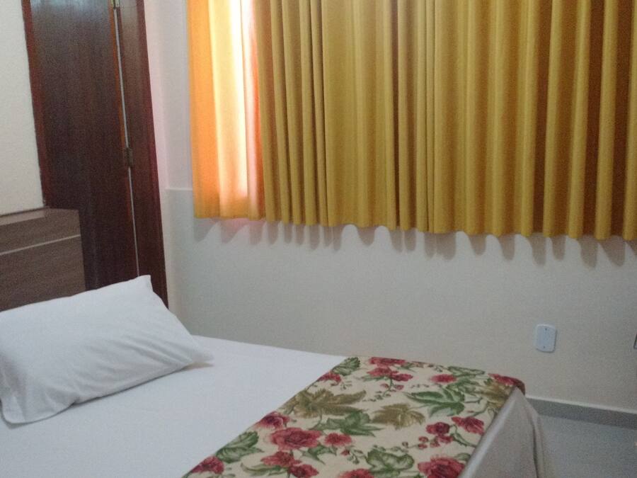Una habitación de hotel con una cama, cabecera de madera, mesita de noche, teléfono y ventana con cortinas amarillas.