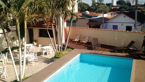 Piscina con sillones y mesas, rodeada de palmeras y un edificio blanco con techo rojo.