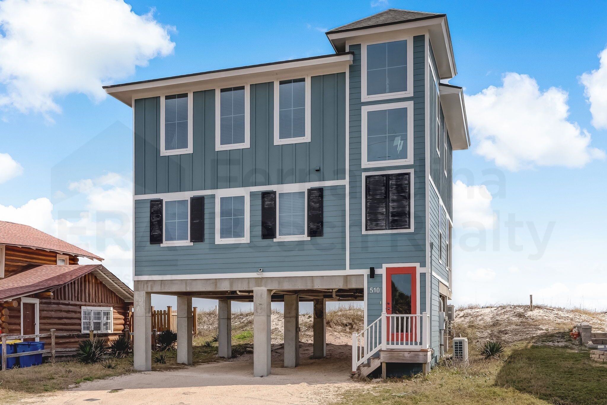 A two-story building with a balcony, a red door, and a sandy ground.