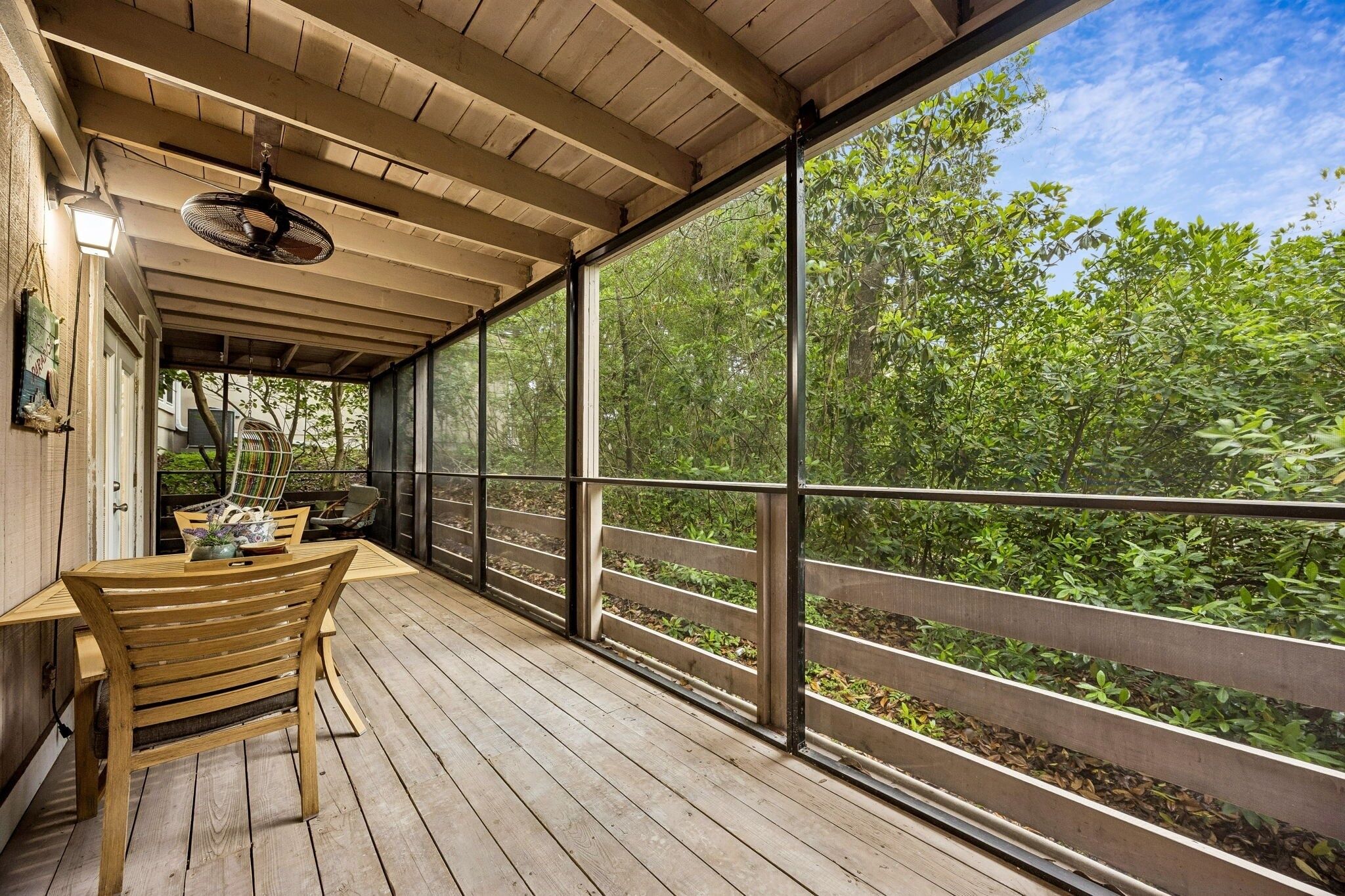 Um deck de madeira com mesa e cadeiras, um ventilador de teto e vista para o verde através de amplas janelas.