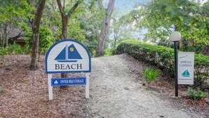 A sign for 'Beach' with a bike storage symbol, surrounded by trees and a gravel path. - Amelia Landings Condo, Easy Access to the Pool and Short Walk to the Beach by RedAwning (Fernandina Beach)