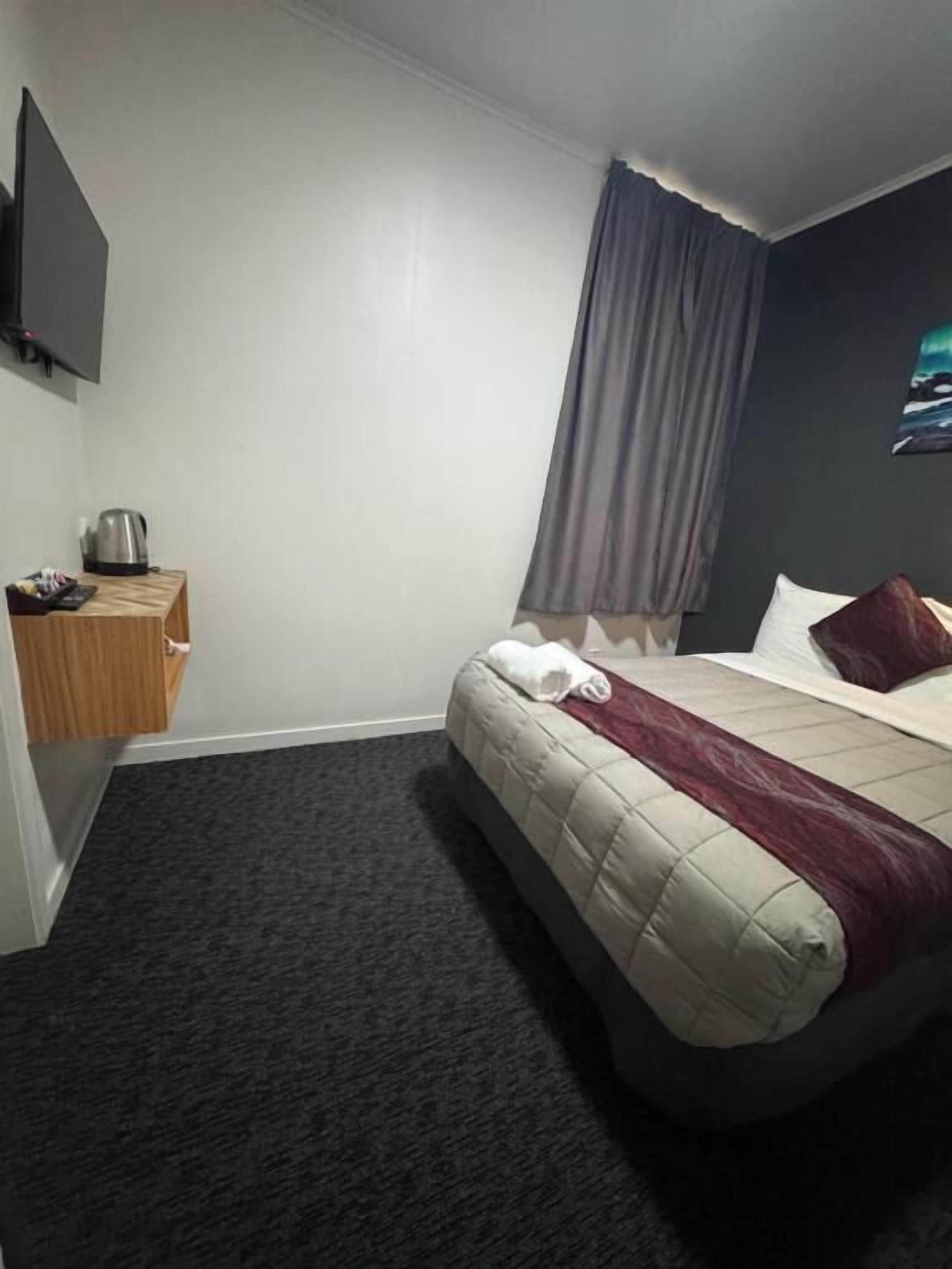 A hotel room with a bed, a small table with a kettle, a flat-screen TV, and a painting on the wall.