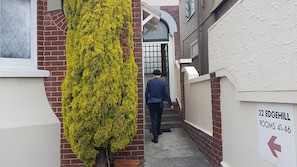 Une ruelle étroite, bordée d’un côté par un mur de briques et de l’autre par une haie verte, mène à une porte dotée d’une fenêtre. Une enseigne indique : « 22 EDGEHILL ROOMS 41-46 ».