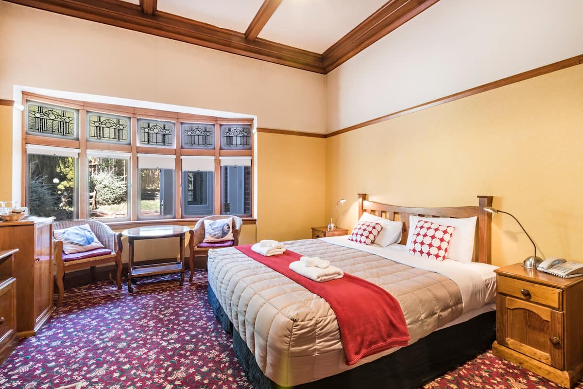 Une chambre d’hôtel avec un grand lit, une table de chevet en bois, un bureau avec une chaise et une fenêtre ornée de vitraux décoratifs.