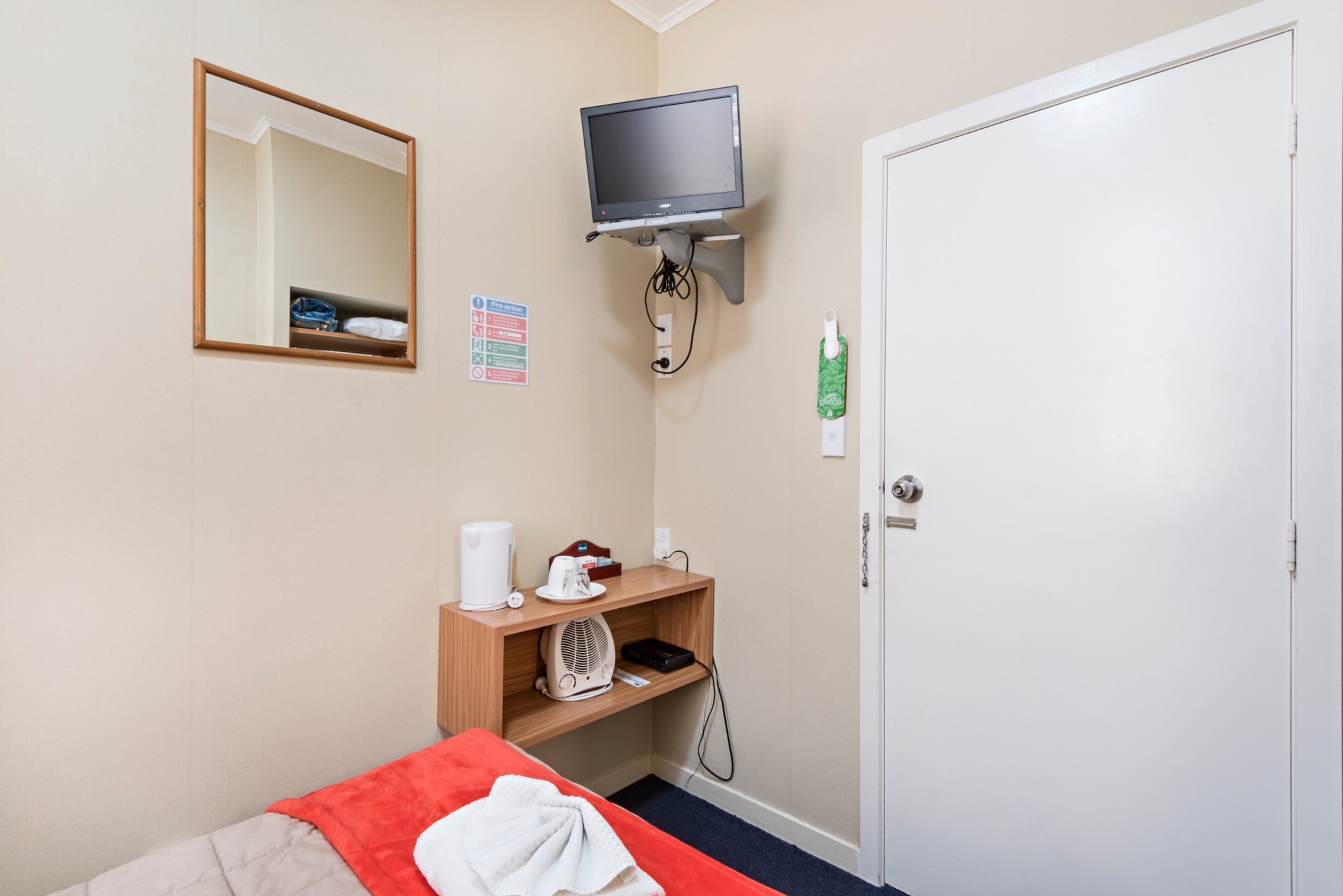 A hotel room with a bed, a television mounted on the wall, a small shelf with a kettle and a fan, and a door with a lock.